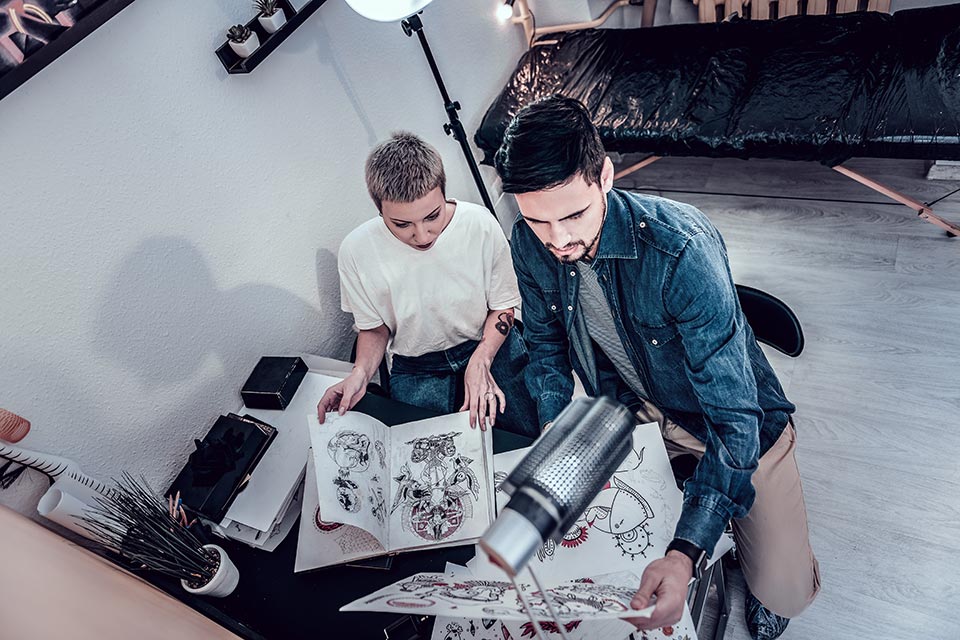 sketches-designs-unusual-tattoo-master-her-male-customer-reaching-interesting-designs-while-sitting-her-office-corner tattoo artist looking at a tattoo apprentice portfolio