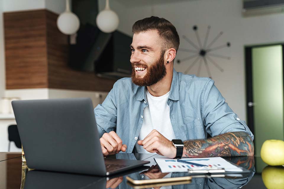smiling-young-bearded-man-working-laptop-computer-while-sitting-kitchen-table__ tattoo shop owner writing a business plan